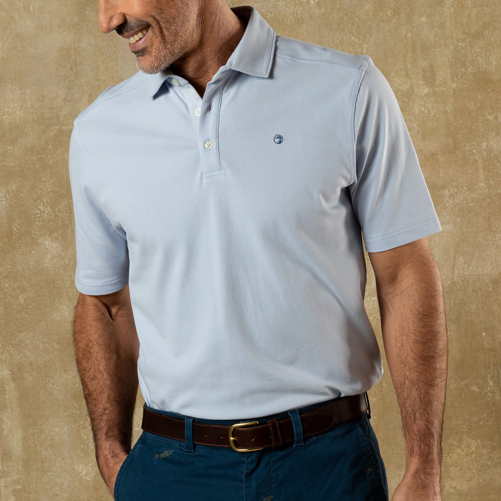 A man stands against a tan textured background wearing Duck Head’s Hanover Pima Cotton Pique Polo in light blue, tucked into blue pants with a brown belt, exemplifying classic style.