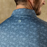 Close-up of a man's back and shoulder wearing the Duck Head Long Drive Performance Polo, a blue golf shirt featuring bird patterns and a small circular Duck Head logo beneath the collar.