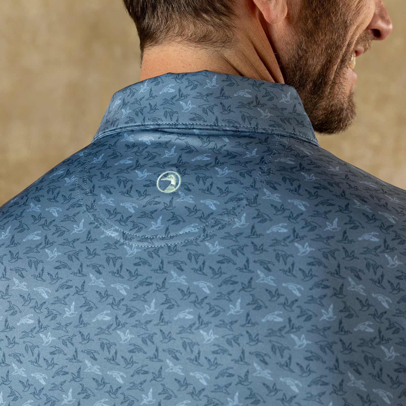 Close-up of a man's back and shoulder wearing the Duck Head Long Drive Performance Polo, a blue golf shirt featuring bird patterns and a small circular Duck Head logo beneath the collar.