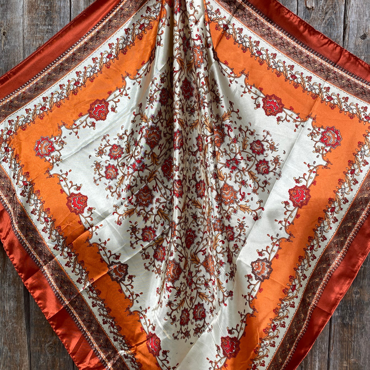 The Burnt Orange and Tan Wild Rag by Rodeo Drive features intricate red and brown floral patterns on a cream background with orange and rust borders, beautifully displayed on a wooden surface.