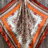 The Burnt Orange and Tan Wild Rag by Rodeo Drive features intricate red and brown floral patterns on a cream background with orange and rust borders, beautifully displayed on a wooden surface.