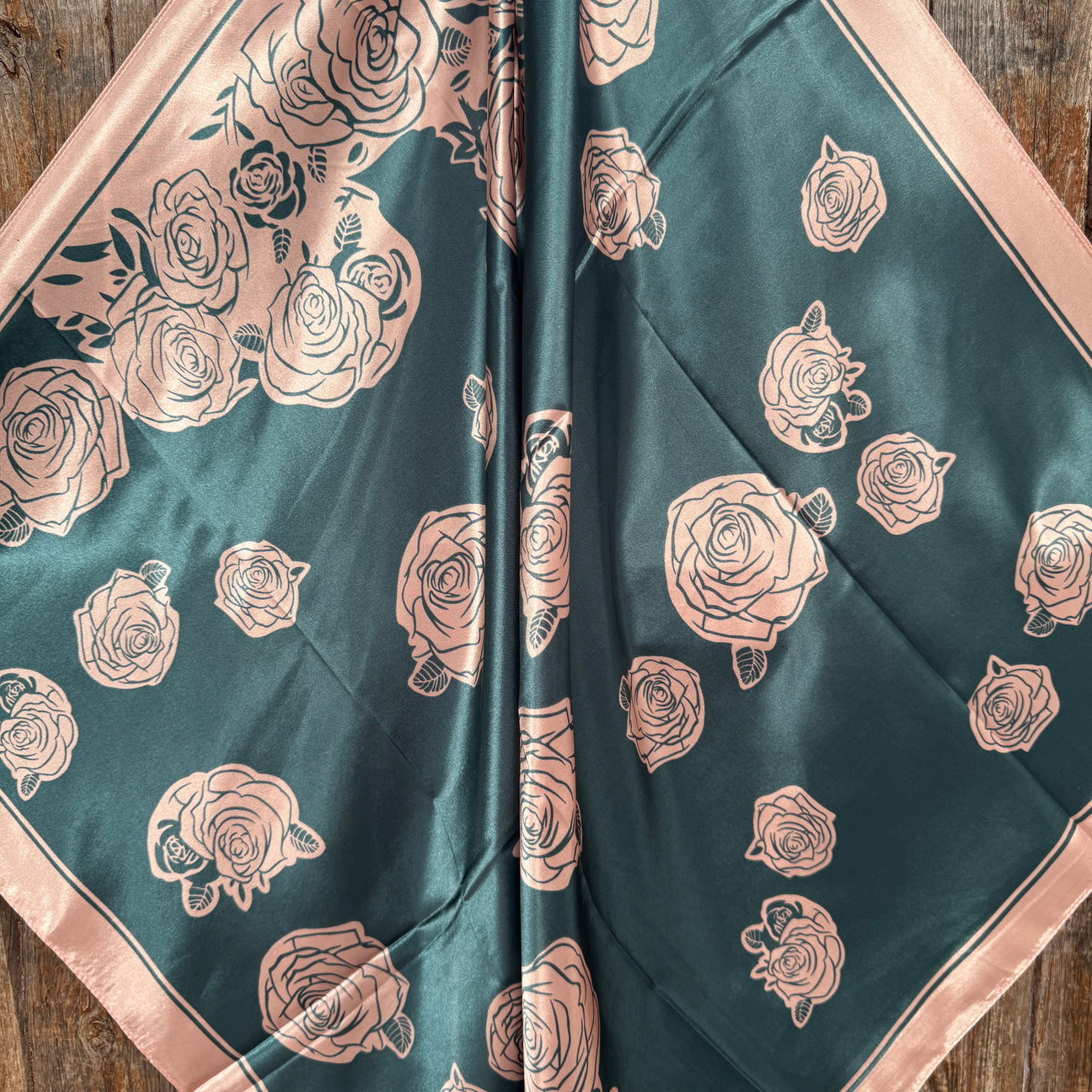 The Baby Pink Roses Wild Rag by Rodeo Drive is a square silk scarf with pink rose patterns on a dark green background and pink border, shown flat on wood—ideal as a chic western accessory or wild rag.