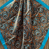 The Blue Floral Wild Rag by Rodeo Drive, featuring a paisley pattern in blue, orange, and brown with a solid blue border, is displayed on a wooden surface.