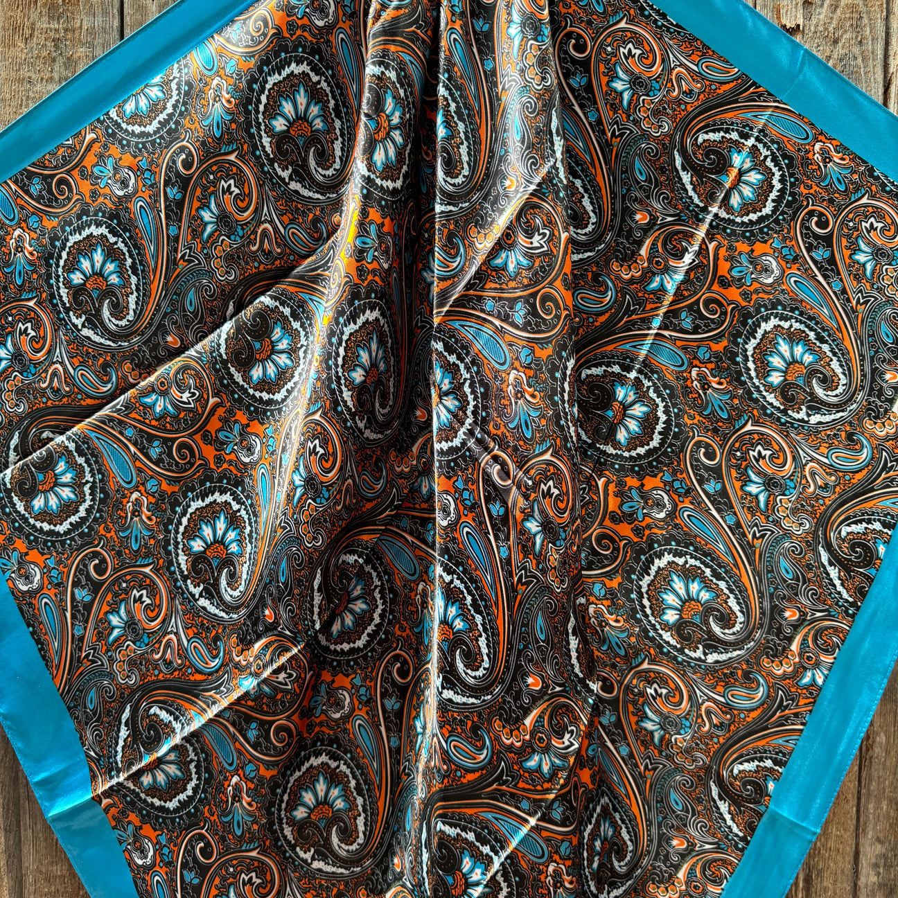 The Blue Floral Wild Rag by Rodeo Drive, featuring a paisley pattern in blue, orange, and brown with a solid blue border, is displayed on a wooden surface.