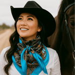 A woman in a black cowboy hat and the Blue & Black Floral Wild Rag by Rodeo Drive stands next to a horse, smiling and looking slightly upwards.
