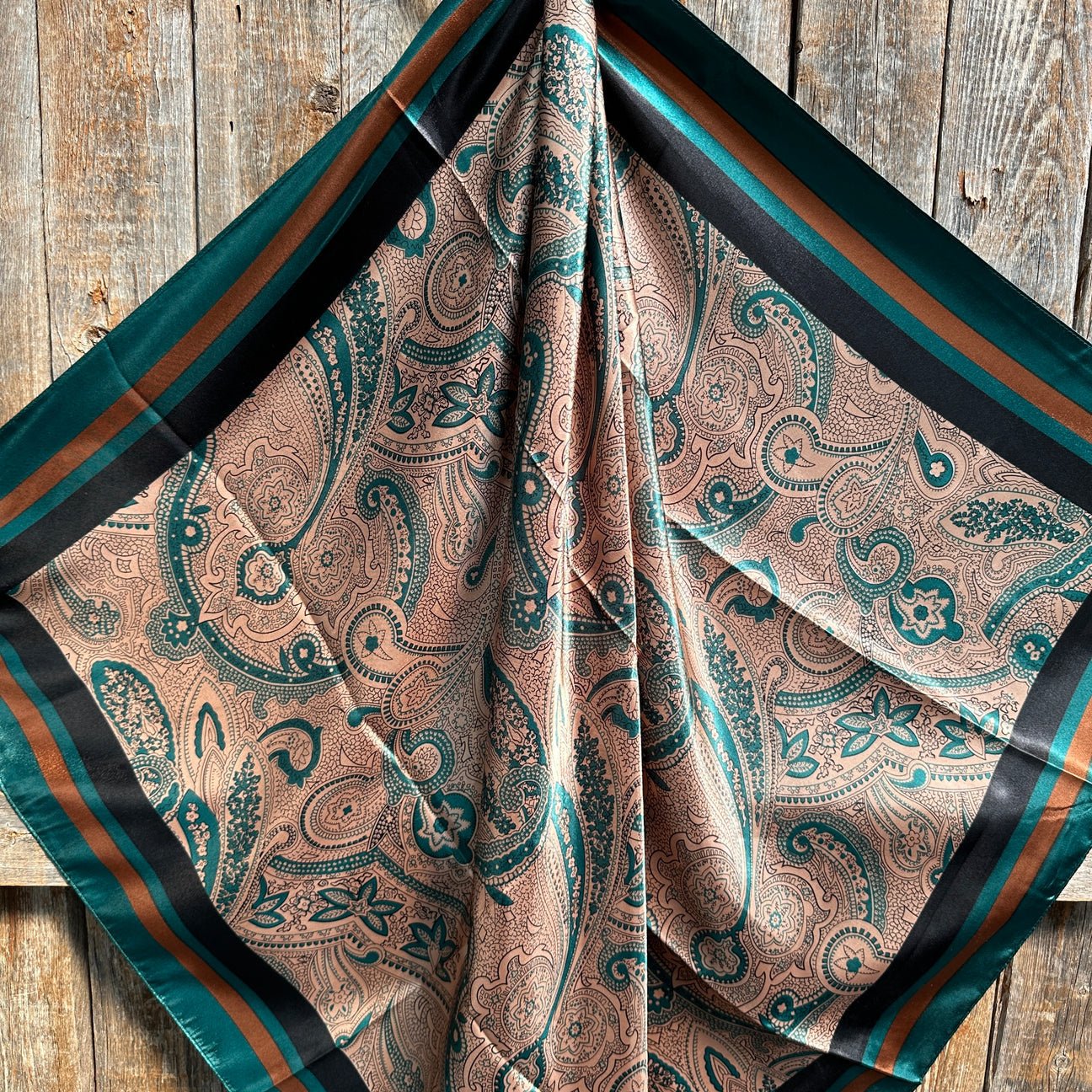 The Emerald and Tan Paisley Wild Rag by Rodeo Drive features green and brown paisley patterns with a striped border, elegantly draped on rustic wood for a classic western touch.