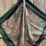 The Emerald and Tan Paisley Wild Rag by Rodeo Drive features green and brown paisley patterns with a striped border, elegantly draped on rustic wood for a classic western touch.