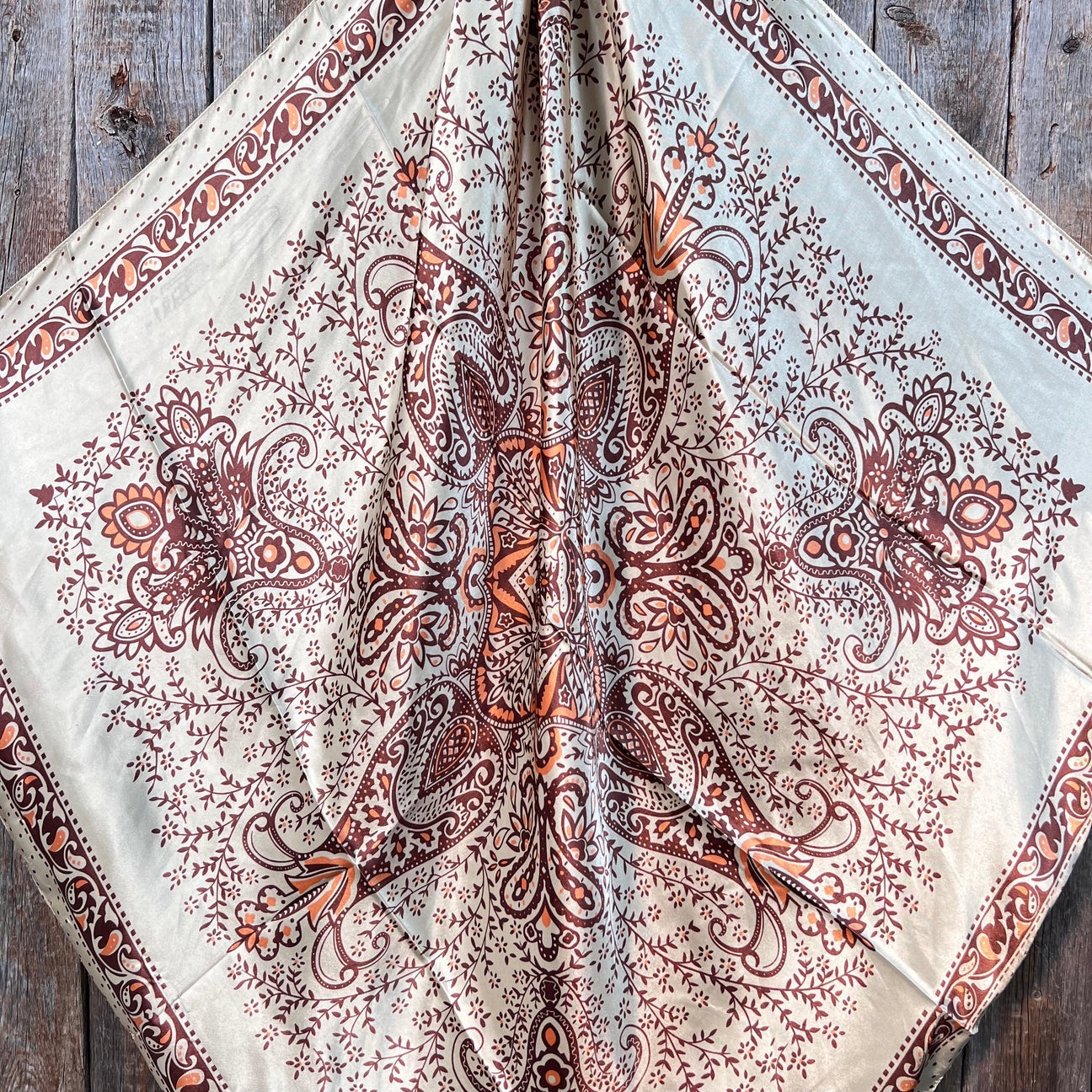 The Tan/Brown Polka Dot and Paisley Wild Rag by Rodeo Drive features an intricate symmetrical pattern with brown and orange motifs on a light background, displayed on a wooden surface.
