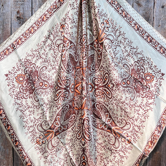 The Tan/Brown Polka Dot and Paisley Wild Rag by Rodeo Drive features an intricate symmetrical pattern with brown and orange motifs on a light background, displayed on a wooden surface.