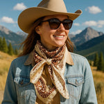 A woman outdoors in the mountains under a blue sky wears a cowboy hat, sunglasses, denim jacket, and the Tan/Brown Polka Dot and Paisley Wild Rag by Rodeo Drive.