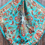 The Rodeo Drive Turquoise That's So 70's Paisley Wild Rag is a square silk scarf with a turquoise base and intricate red, green, and beige paisley floral patterns, displayed on a wooden surface.