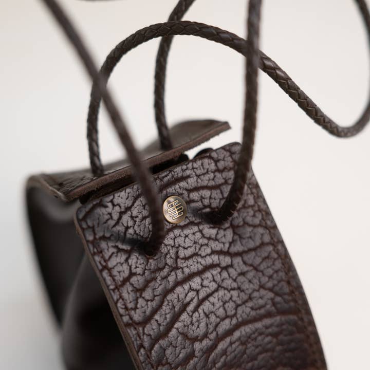 Close-up of the Joi Bag in Coffee by Stash—a textured dark brown leather drawstring bag with braided straps and a small round metal stud engraved with the Stash logo, highlighting its subtle Japanese-inspired handbag details.