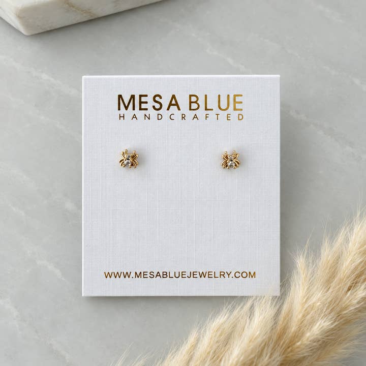 A pair of Tiny Cz Spider Stud Earrings by Mesa Blue, featuring 18K gold plating and pave CZ accents, displayed on a white Mesa Blue jewelry card placed on a marble surface.