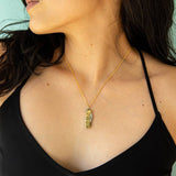 A woman wearing a black spaghetti strap top and the Ornamental Things Gold Wildflower Knife Necklace, featuring a rectangular pendant, faces sideways against a light-colored background.