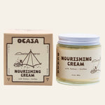 A square box and a glass jar of OCASA Nourishing Coffee-Infused Tallow Cream, both featuring illustrations of a tent and coffee brewing with labels reading “Nourishing Cream with Tallow + Coffee.”