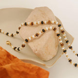 Arrow Boho Beaded Necklace by Flanerie by MAIQ featuring white, brown, and black hypoallergenic beads with 18K gold-tone spacers, draped over a decorative stone.