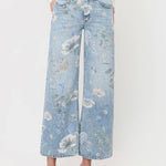 A person styled in Vervet’s Super High Rise Floral Extreme Vintage Flare Jeans, featuring a light blue floral print and dramatic wide-leg silhouette, paired with a white long-sleeve top and white strappy sandals, standing against a plain light background.