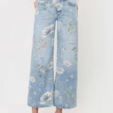 A person styled in Vervet’s Super High Rise Floral Extreme Vintage Flare Jeans, featuring a light blue floral print and dramatic wide-leg silhouette, paired with a white long-sleeve top and white strappy sandals, standing against a plain light background.