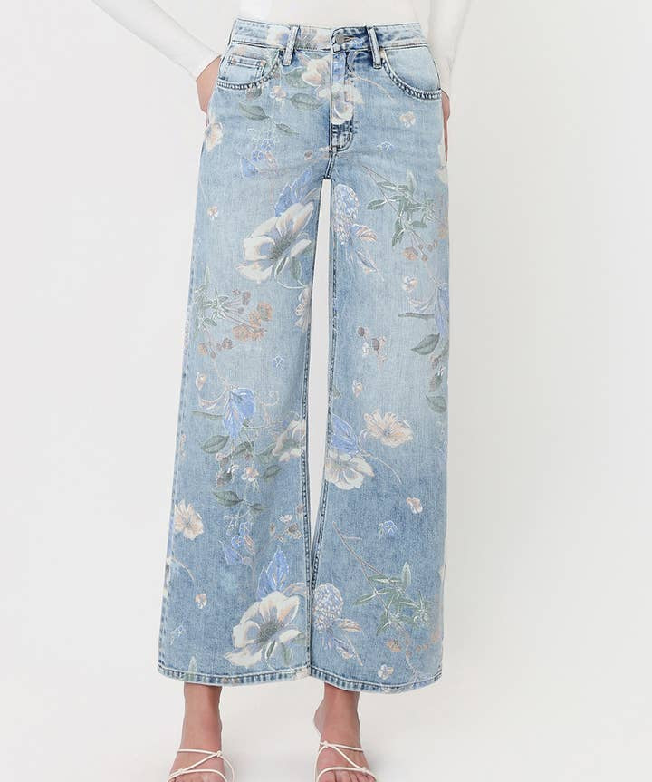 A person styled in Vervet’s Super High Rise Floral Extreme Vintage Flare Jeans, featuring a light blue floral print and dramatic wide-leg silhouette, paired with a white long-sleeve top and white strappy sandals, standing against a plain light background.