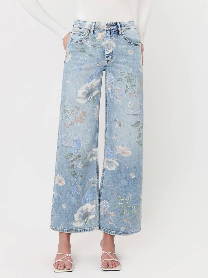 A person styled in Vervet’s Super High Rise Floral Extreme Vintage Flare Jeans, featuring a light blue floral print and dramatic wide-leg silhouette, paired with a white long-sleeve top and white strappy sandals, standing against a plain light background.