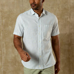 A man wears the Duck Head Linen Cotton Oxford Sport Shirt, featuring light blue and white vertical stripes, a button-down collar, and front pocket, standing against a tan textured background.