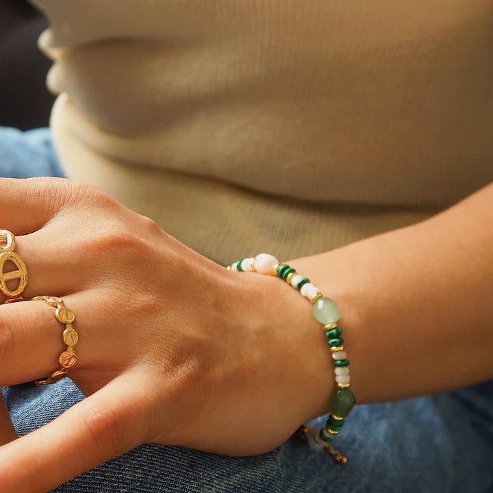 A Faye Bohemian Stone and Bead Bracelet with alternating green, white, and gold-toned beads on a wrist, worn with two 18K gold plated chain rings.