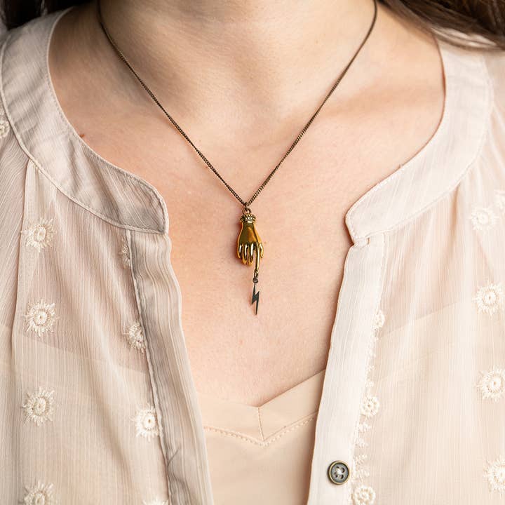 Magician’s Hand Necklace – Aged Brass Chain featuring an antique gold hand holding a small lightning bolt pendant on a thin chain.