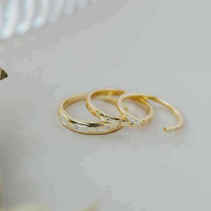 Three Gold Rings | Inset Baguette by JaxKelly are arranged in a row on a light surface, with part of a textured glass object visible in the background.