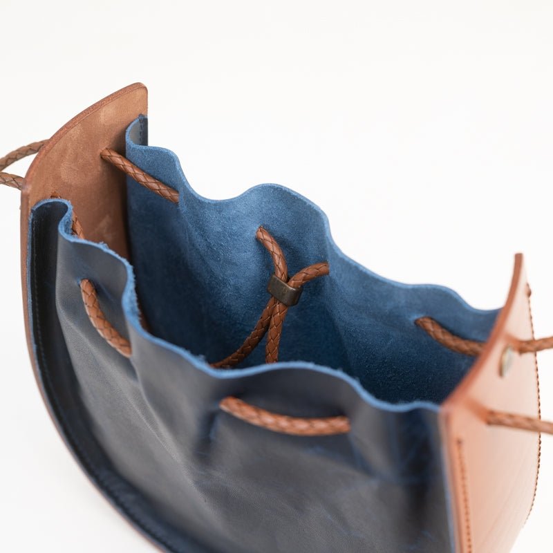 The Joi Bag | Navy by Stash features an open leather drawstring design with a blue suede interior, brown leather exterior panels, and a braided drawstring closure, viewed from above. This Japanese-inspired bag combines unique style and functionality in its distinctive design.
