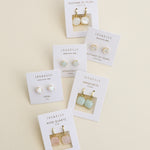 Six pairs of JaxKelly Gemstone Mini Drop earrings on display cards are arranged on a beige background, featuring mother of pearl, opal, amazonite, and Rose Quartz designs. These stylish and durable pieces from JaxKelly are tarnish-resistant—perfect for everyday wear.