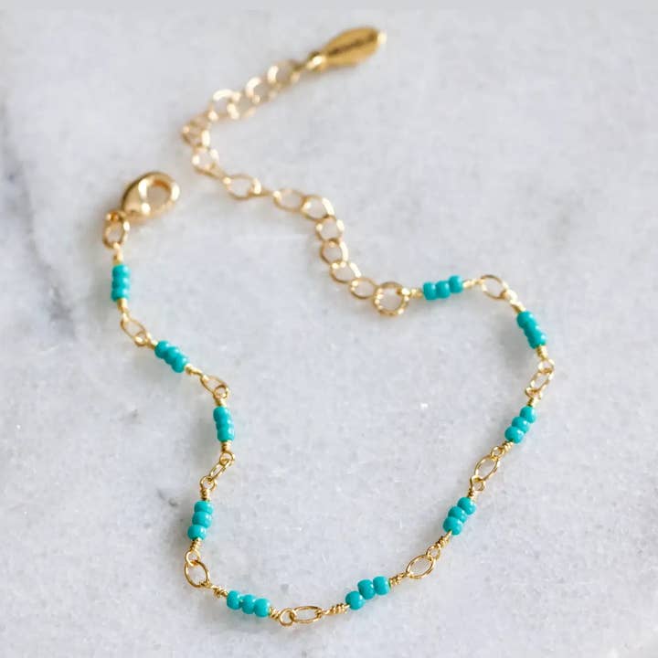 Mesa Blue’s Turquoise Seed Bead Bracelet features small turquoise beads evenly spaced along an 18K gold plated chain, complete with a lobster clasp and adjustable extender. Shown here on a light surface.