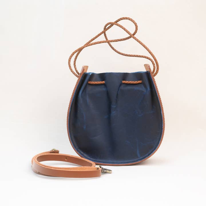 The Joi Bag in Navy by Stash is a small leather crossbody featuring brown trim and a braided strap, inspired by Japanese design aesthetics. It is displayed with an additional detachable tan strap placed in front of the bag.
