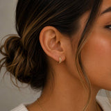 A close-up of a woman’s ear wearing the Double Champagne Hoop by JaxKelly—a small gold double hoop earring with her brown hair styled in a loose bun and a strand softly framing her face. One delicate hoop is embellished with shimmering pave stones, adding subtle sparkle to this hypoallergenic accessory from JaxKelly.
