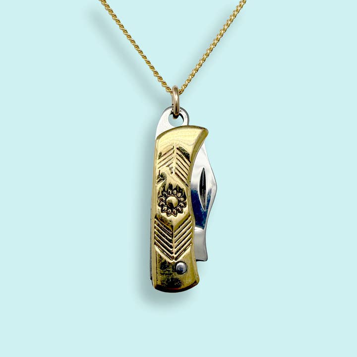 The Gold Wildflower Knife Necklace by Ornamental Things features a functional gold and silver knife pendant suspended from a gold chain, displayed against a light blue background.