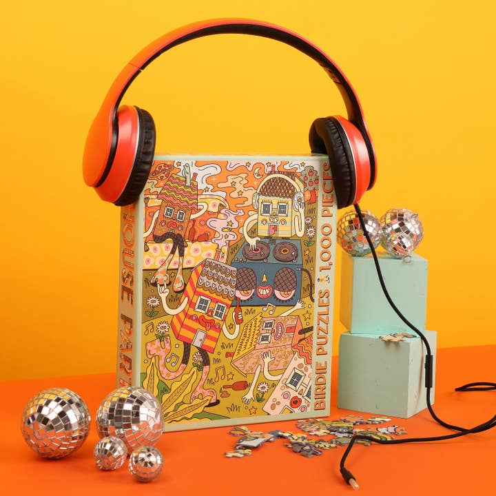 The House Party 1,000 Piece Puzzle by Birdie Puzzles features a vibrant recycled box with orange headphones on top, surrounded by disco balls and puzzle pieces, set against a yellow-orange background.