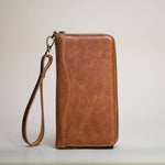 The Stash Passport Wallet in Caramel is displayed upright on a plain surface against a neutral background, featuring a matching wrist strap and zippered design—an ideal choice for those seeking a stylish leather travel wallet.