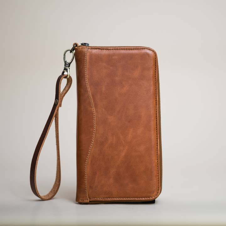 The Stash Passport Wallet in Caramel is displayed upright on a plain surface against a neutral background, featuring a matching wrist strap and zippered design—an ideal choice for those seeking a stylish leather travel wallet.