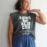 A woman with curly hair stands against a plain wall, looking to the side, wearing a Ramble & Company boxy fit tee featuring the “Doing My Best Club” design and blue plaid shorts.