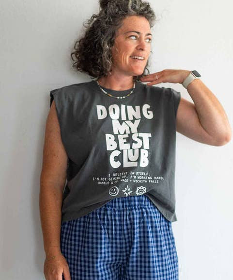 A woman with curly hair stands against a plain wall, looking to the side, wearing a Ramble & Company boxy fit tee featuring the “Doing My Best Club” design and blue plaid shorts.