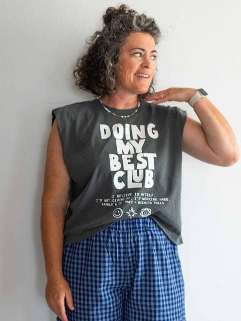 A woman with curly hair stands against a plain wall, looking to the side, wearing a Ramble & Company boxy fit tee featuring the “Doing My Best Club” design and blue plaid shorts.