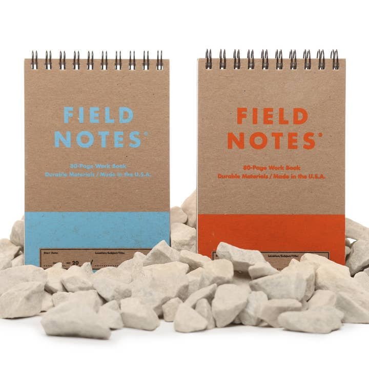 Two spiral-bound Field Notes 80-Page Work Books, one with blue text and one with orange, stand upright among white rocks. Their covers feature Super Duty Chipboard for added durability. Brand: Field Notes.
