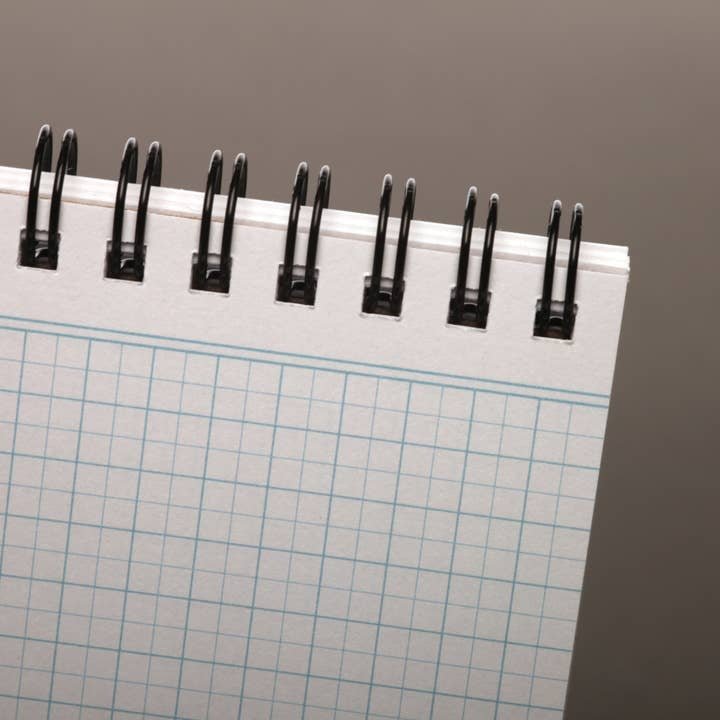 A close-up of the top corner of a spiral-bound 80-Page Work Book by Field Notes with graph paper, wire binding, Double Graph Grid, and a grey background.
