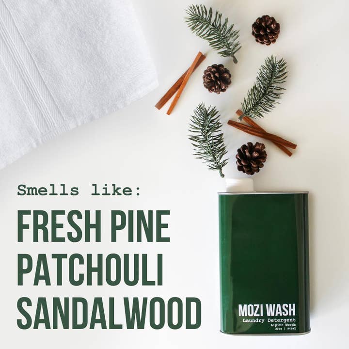A bottle of Mozi Alpine Woods Laundry Detergent sits beside a folded white towel, surrounded by pine branches, pinecones, and cinnamon sticks; text reads “Smells like: Fresh pine, patchouli, sandalwood.”.