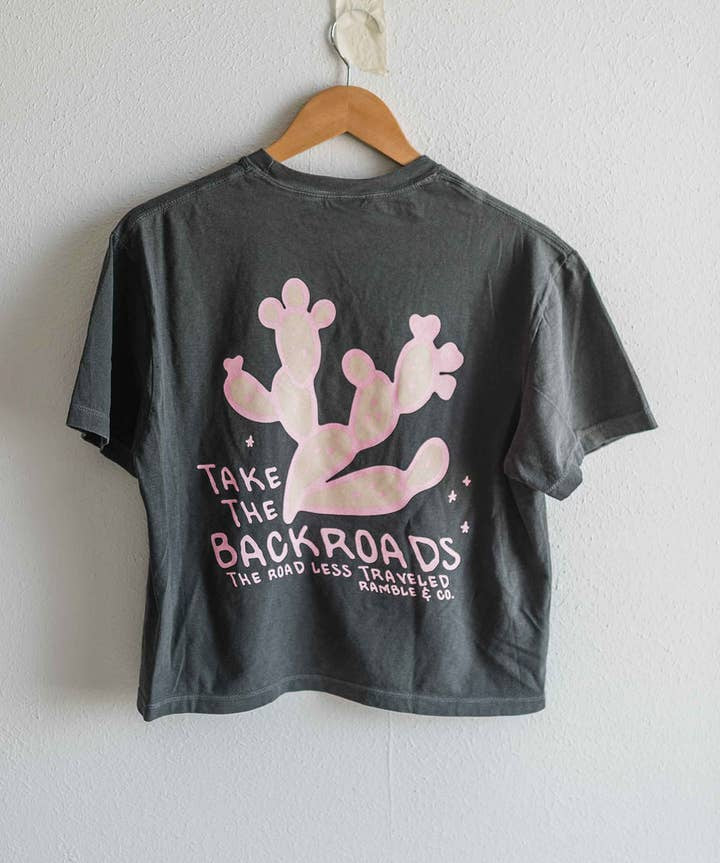 The Take the Backroads Crop from Ramble & Company is a dark gray, boxy-fit t-shirt displayed on a hanger, featuring a pink cactus graphic and the text “Take the Backroads, The Road Less Traveled, Ramble & Company” across the back. This Comfort Colors crop tee is ideal for those who want to slow down, make memories, and embrace adventure in style.