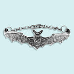 The Silver Bat Bracelet by Ornamental Things showcases a detailed, vintage-inspired bat with outstretched wings, attached to a chain and clasp. This striking piece of gothic jewelry is displayed on a light blue background.