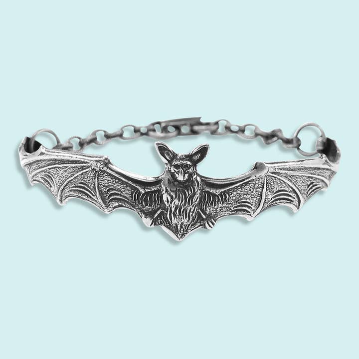 The Silver Bat Bracelet by Ornamental Things showcases a detailed, vintage-inspired bat with outstretched wings, attached to a chain and clasp. This striking piece of gothic jewelry is displayed on a light blue background.