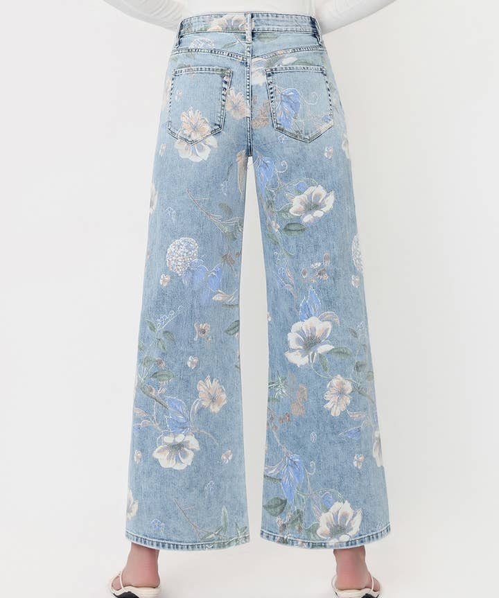 A person is shown from the back against a plain white background, wearing the Vervet Super High Rise Floral Extreme Vintage Flare Jeans in light blue with a wide-leg silhouette and white heeled shoes.
