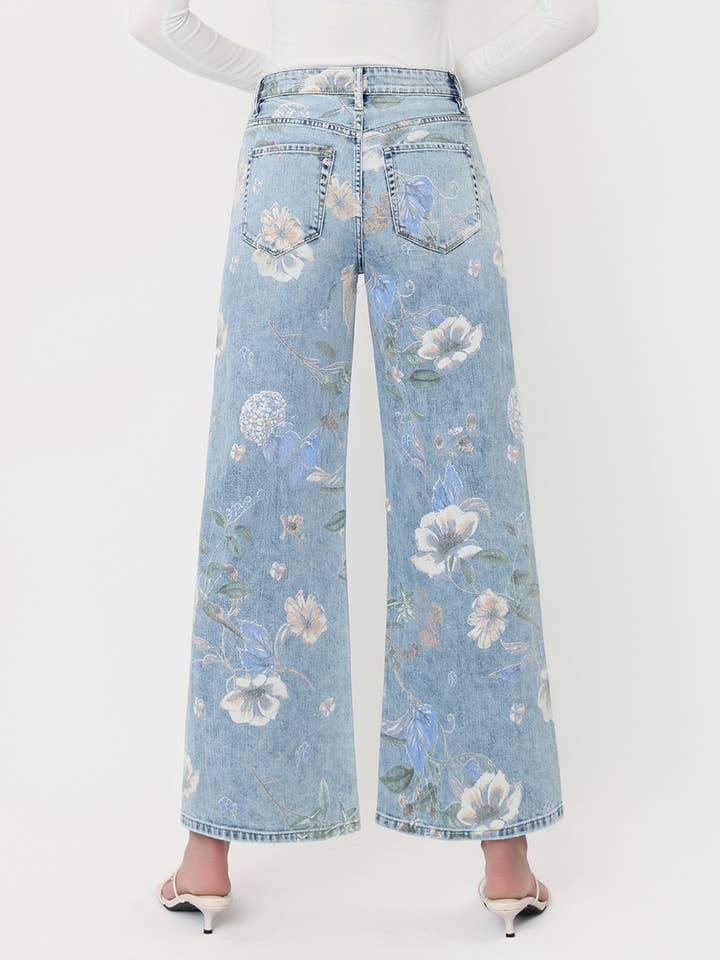 A person is shown from the back against a plain white background, wearing the Vervet Super High Rise Floral Extreme Vintage Flare Jeans in light blue with a wide-leg silhouette and white heeled shoes.