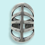 The Silver Gladiator Ring by Ornamental Things features a wide, silver-toned metal band with an openwork design of horizontal and vertical cut-outs, reminiscent of gladiator sandals, elegantly displayed on a light blue background.