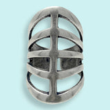 The Silver Gladiator Ring by Ornamental Things features a wide, silver-toned metal band with an openwork design of horizontal and vertical cut-outs, reminiscent of gladiator sandals, elegantly displayed on a light blue background.